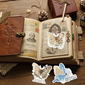May include: An open brown leather journal with a cross embossed on the cover, featuring vintage-style pages with printed portraits, text, and a stained glass illustration. Three butterfly-shaped stickers with text overlays are displayed.