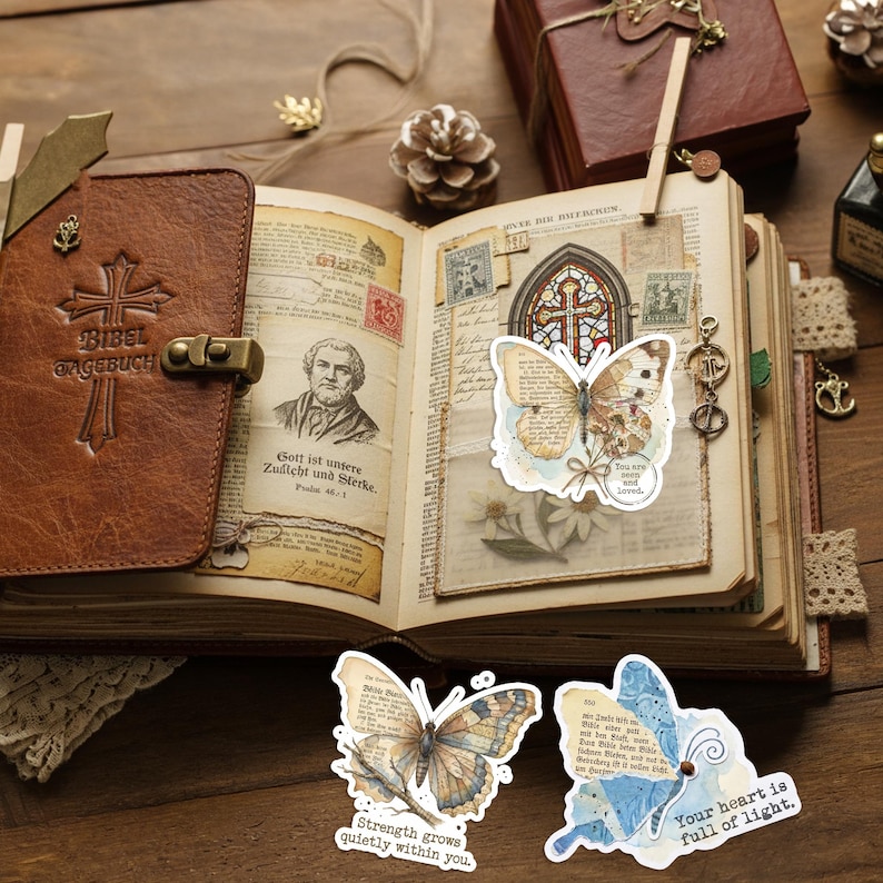 May include: An open brown leather journal with a cross embossed on the cover, featuring vintage-style pages with printed portraits, text, and a stained glass illustration. Three butterfly-shaped stickers with text overlays are displayed.