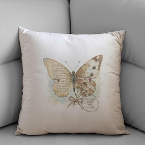 May include: A square off-white throw pillow featuring a central graphic of a butterfly with wings composed of vintage book pages. Below the butterfly is a small tied bow and a circular stamp containing the text, You are seen and loved.