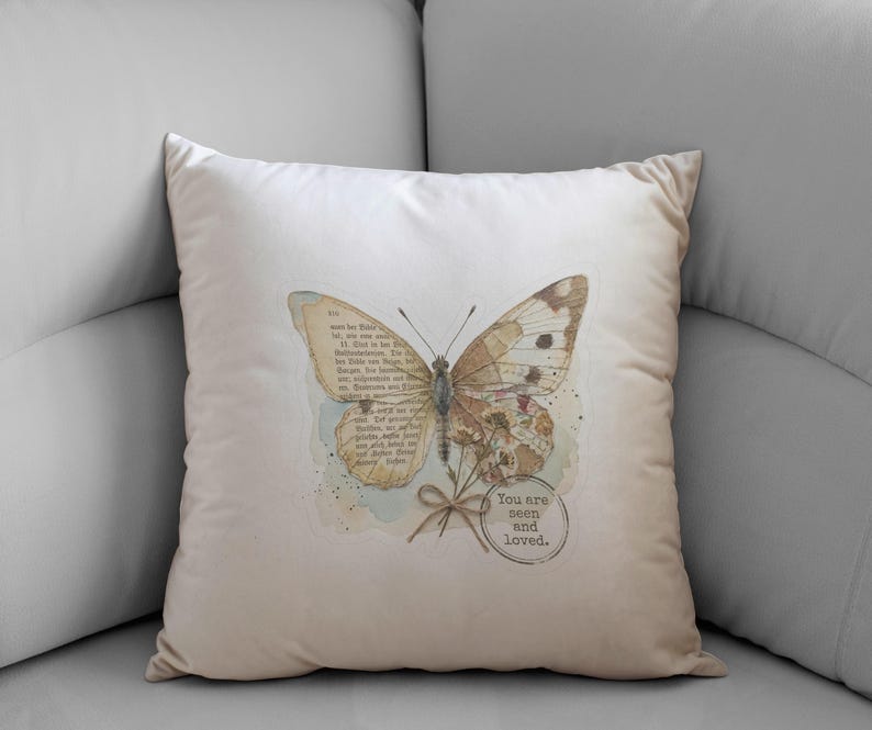 May include: A square off-white throw pillow featuring a central graphic of a butterfly with wings composed of vintage book pages. Below the butterfly is a small tied bow and a circular stamp containing the text, You are seen and loved.