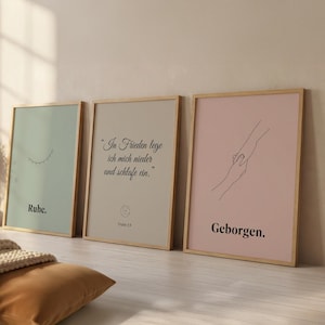 May include: Three framed art prints with wooden frames. The prints feature minimalist designs and text in German. One print is light green with a closed eye and the word "Ruhe." Another is beige with text and a symbol. The third is pink with hands and the word "Geborgen."