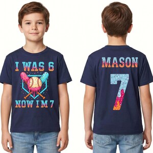 May include: Navy blue short-sleeve t-shirt with a baseball-themed graphic. The front of the shirt says "I WAS 6 NOW I'M 7". The back of the shirt has the name "MASON" and the number "7".