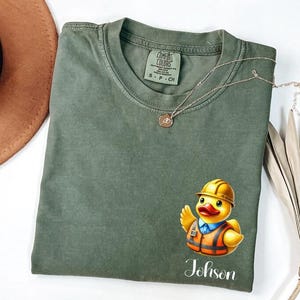 May include: An olive green crew neck t-shirt featuring a graphic of a yellow rubber duck wearing a construction hard hat and an orange safety vest. Below the duck, the text reads "Johnson" in a white script font. The shirt has a Comfort Colors label.