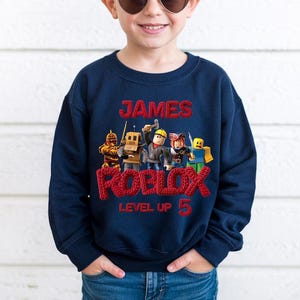 Puede incluir: Sudadera azul marino con el nombre "JAMES" en rojo, encima de "ROBLOX" y "LEVEL UP 5". El dise&ntilde;o incluye gr&aacute;ficos de personajes de Roblox. La sudadera la lleva una persona.