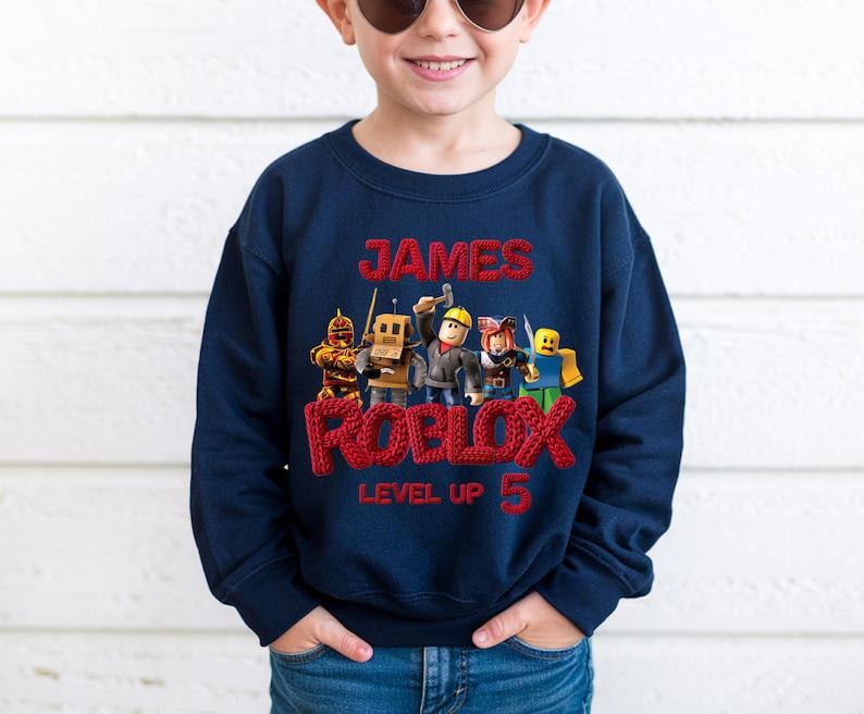 Puede incluir: Sudadera azul marino con el nombre "JAMES" en rojo, encima de "ROBLOX" y "LEVEL UP 5". El dise&ntilde;o incluye gr&aacute;ficos de personajes de Roblox. La sudadera la lleva una persona.