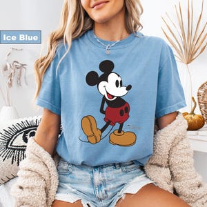 May include: Ice blue t-shirt featuring a classic Mickey Mouse graphic. The cartoon character is depicted in black, red, and tan colors. The shirt has short sleeves and a crew neck.