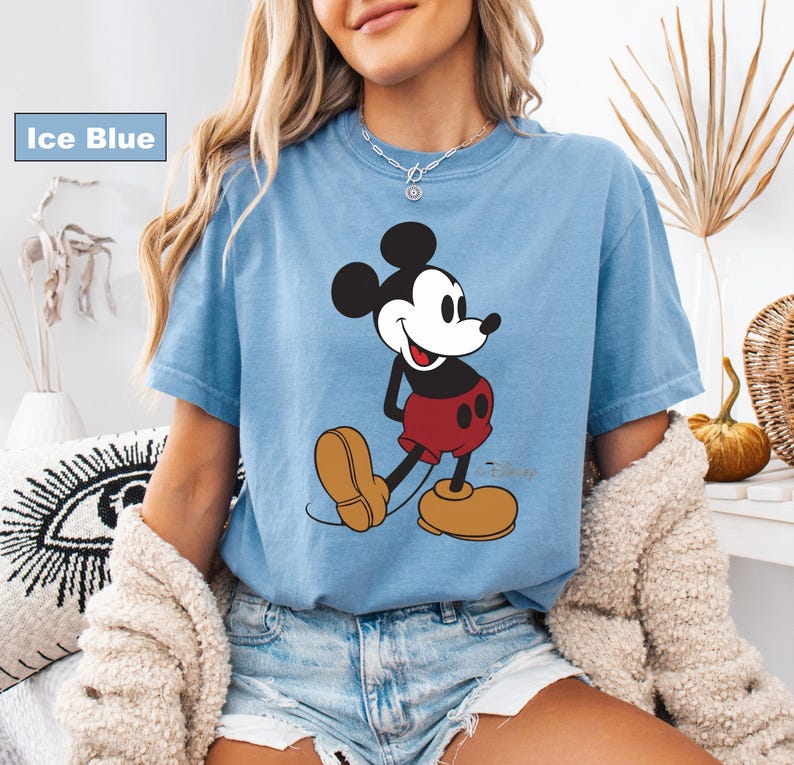 May include: Ice blue t-shirt featuring a classic Mickey Mouse graphic. The cartoon character is depicted in black, red, and tan colors. The shirt has short sleeves and a crew neck.