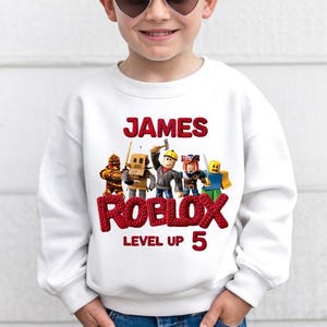 Puede incluir: Sudadera blanca con el nombre "JAMES" en rojo, encima de "ROBLOX" y "LEVEL UP 5". El dise&ntilde;o incluye personajes de Roblox. Una prenda casual y c&oacute;moda para ni&ntilde;os.
