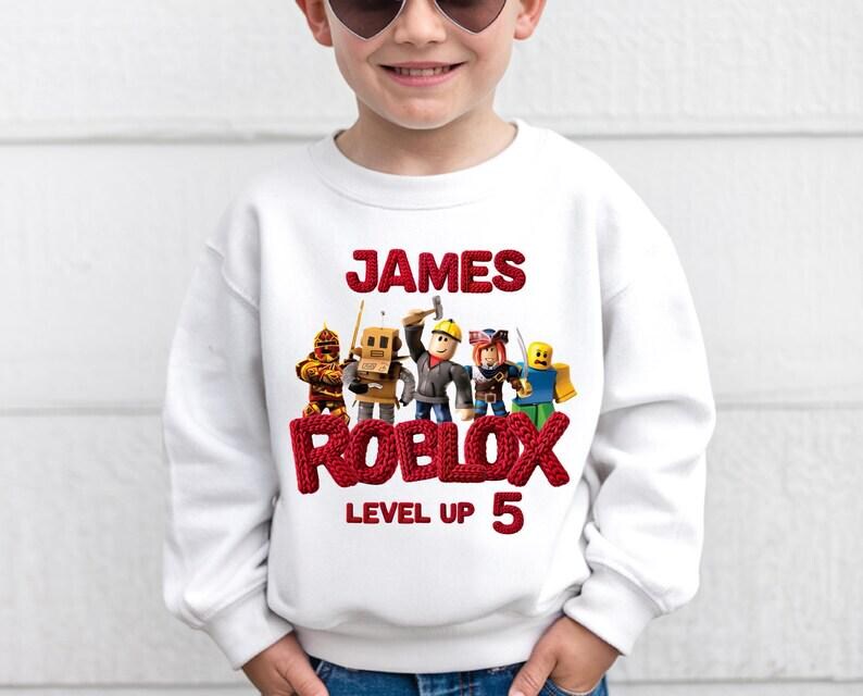 Puede incluir: Sudadera blanca con el nombre "JAMES" en rojo, encima de "ROBLOX" y "LEVEL UP 5". El dise&ntilde;o incluye personajes de Roblox. Una prenda casual y c&oacute;moda para ni&ntilde;os.