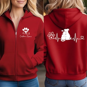 May include: A red zip-up hoodie featuring a white graphic on the front left chest of a paw print with a heart and the text Custom Name. The back displays a white graphic of a heartbeat line, a paw print, and a cat and dog hugging with a heart.