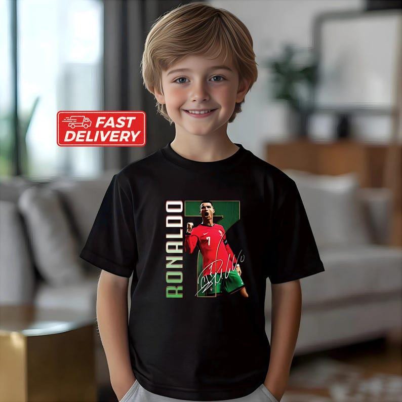Puede incluir: Camiseta negra con un gr&aacute;fico de un futbolista con camiseta roja, pantal&oacute;n corto verde y firma. La palabra "RONALDO" est&aacute; impresa verticalmente en verde y blanco. Un ni&ntilde;o lleva la camiseta. La imagen tambi&eacute;n incluye una pegatina de "Entrega r&aacute;pida".