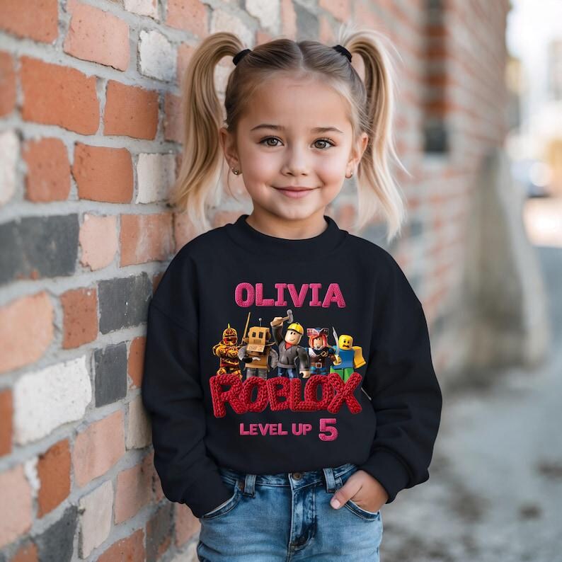 Puede incluir: Una sudadera negra para ni&ntilde;os con el nombre "OLIVIA" en rosa, la palabra "ROBLOX" y la frase "LEVEL UP 5". La sudadera presenta un gr&aacute;fico colorido de personajes de Roblox.