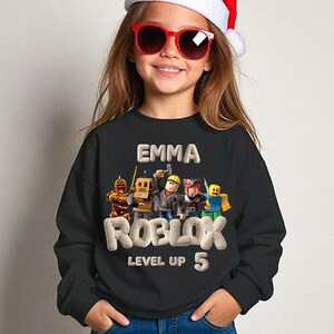Puede incluir: Sudadera negra con el nombre "EMMA" y la palabra "ROBLOX" en una fuente texturizada, junto con "LEVEL UP 5". El dise&ntilde;o incluye personajes de dibujos animados. La sudadera la lleva una persona con gafas de sol rojas y un gorro de Pap&aacute; Noel.
