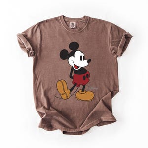 May include: Brown t-shirt featuring a classic Mickey Mouse graphic. The cartoon character is depicted in black, white, red, and yellow. The Disney logo is visible at the bottom right of the graphic.