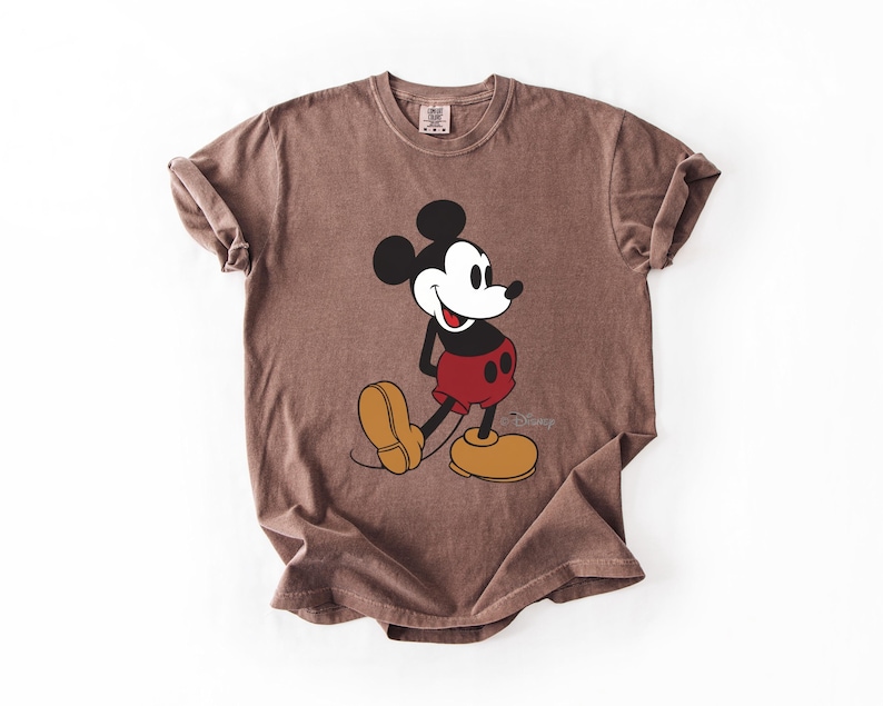 May include: Brown t-shirt featuring a classic Mickey Mouse graphic. The cartoon character is depicted in black, white, red, and yellow. The Disney logo is visible at the bottom right of the graphic.