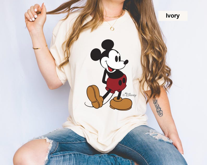 May include: Ivory-colored t-shirt featuring a classic Mickey Mouse graphic. The cartoon character is depicted in black, red, and brown, with the Disney logo visible. The shirt has short sleeves and a relaxed fit.