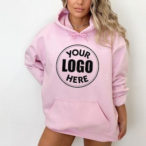 May include: A light pink hooded sweatshirt featuring a large black circular graphic on the chest. Inside the circle, the text reads YOUR LOGO HERE in bold black capital letters. The garment includes a front kangaroo pocket and matching drawstrings.