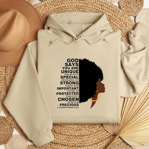 May include: A beige hoodie with a hood and long sleeves. The front features a graphic of a woman's silhouette with an afro and gold hoop earring, and text that reads "GOD SAYS YOU ARE UNIQUE" and other inspirational words.