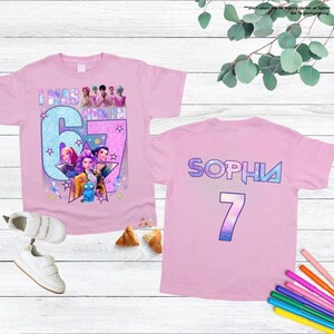 May include: Pink t-shirts with a colorful graphic design. One shirt features the text "I WAS NOMIN 67" with cartoon characters. The other shirt has the name "SOPHIA" and the number "7".