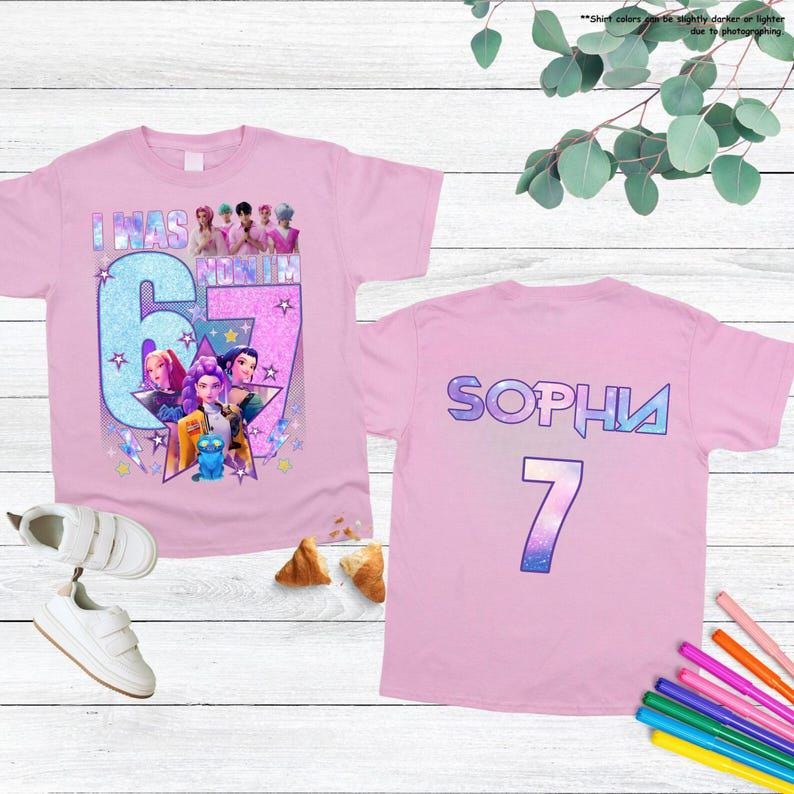 May include: Pink t-shirts with a colorful graphic design. One shirt features the text "I WAS NOMIN 67" with cartoon characters. The other shirt has the name "SOPHIA" and the number "7".