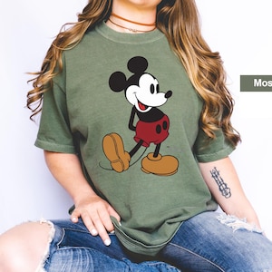 May include: Moss green t-shirt featuring a classic Mickey Mouse graphic. The cartoon character is depicted in black, white, red, and tan. The shirt has short sleeves and a relaxed fit, perfect for casual wear.