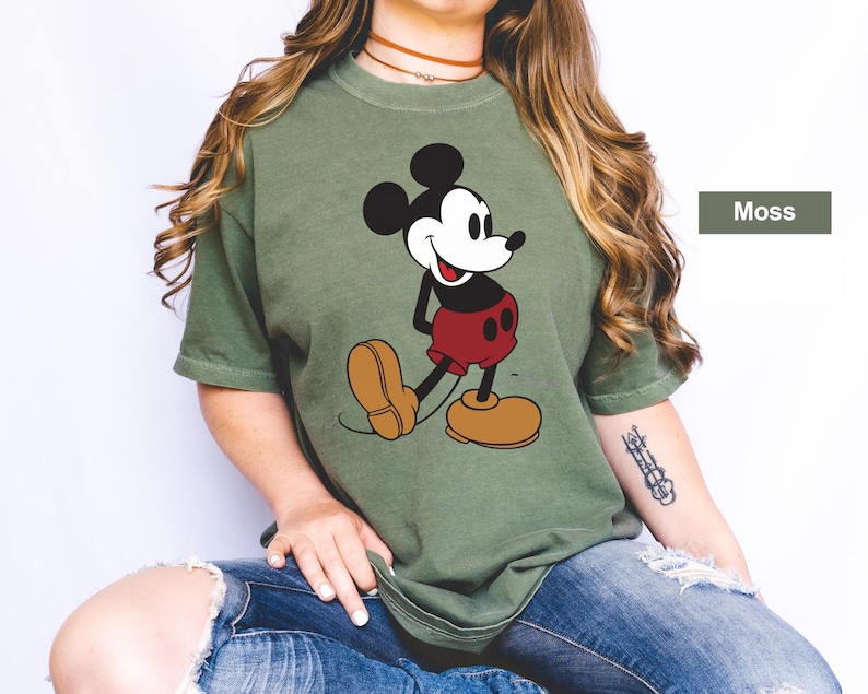 May include: Moss green t-shirt featuring a classic Mickey Mouse graphic. The cartoon character is depicted in black, white, red, and tan. The shirt has short sleeves and a relaxed fit, perfect for casual wear.