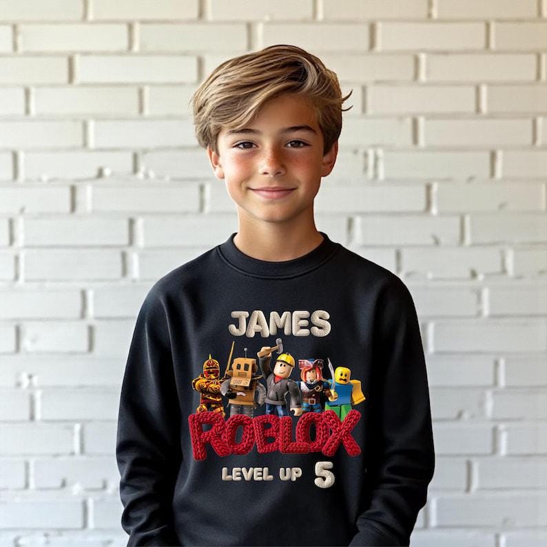 Puede incluir: Sudadera negra con el nombre "JAMES" en blanco y la palabra "ROBLOX" en rojo, con "LEVEL UP 5" en blanco. La sudadera presenta un grupo de personajes de Roblox. La persona de la imagen lleva la sudadera.
