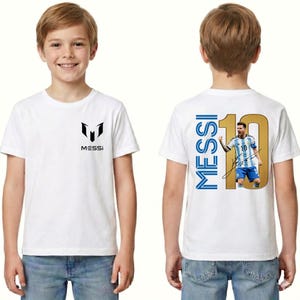 Może przedstawiać: Biała koszulka z napisem "MESSI" i logo z przodu. Tył koszulki przedstawia złoty numer 10 ze zdjęciem piłkarza i napisem "MESSI" w kolorze niebieskim.
