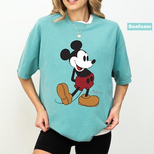 May include: Seafoam green t-shirt featuring a classic Mickey Mouse graphic. The cartoon character is depicted in his signature red shorts, yellow shoes, and black gloves. The shirt has a relaxed fit and a white crew neck.