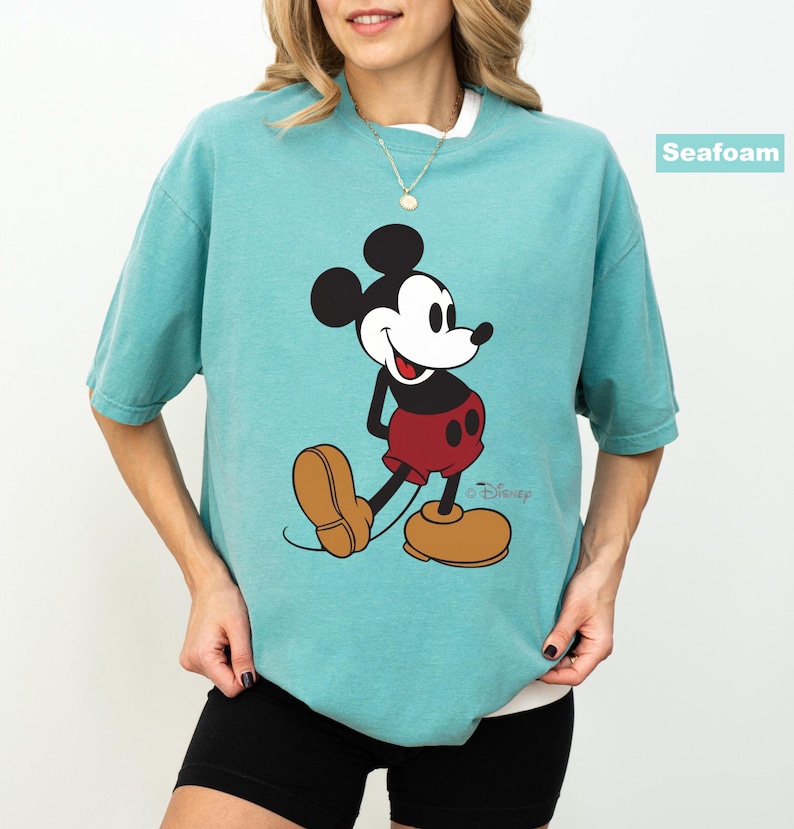 May include: Seafoam green t-shirt featuring a classic Mickey Mouse graphic. The cartoon character is depicted in his signature red shorts, yellow shoes, and black gloves. The shirt has a relaxed fit and a white crew neck.