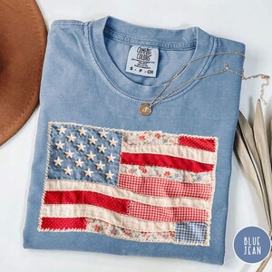 May include: A blue short-sleeved t-shirt featuring a rectangular patch of an American flag made from various fabric patterns including red polka dots, floral prints, and gingham. The flag includes white embroidered stars and a Comfort Colors label.
