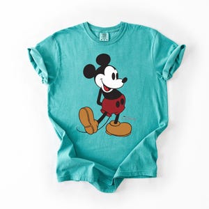 May include: A turquoise t-shirt featuring a graphic of Mickey Mouse. The cartoon character is depicted in his classic pose, wearing red shorts, yellow shoes, and black gloves. The shirt is made of a soft material and has a relaxed fit.