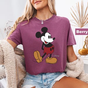 May include: Berry-colored t-shirt featuring a graphic of Mickey Mouse. The cartoon character is depicted in his classic outfit: red shorts, yellow shoes, and black gloves. The shirt is a relaxed fit.