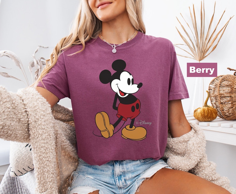 May include: Berry-colored t-shirt featuring a graphic of Mickey Mouse. The cartoon character is depicted in his classic outfit: red shorts, yellow shoes, and black gloves. The shirt is a relaxed fit.