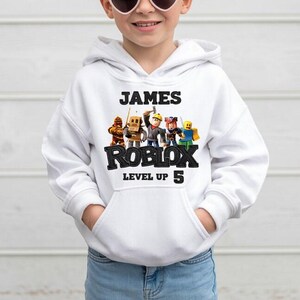 Puede incluir: Sudadera con capucha blanca con el nombre "JAMES" encima de "ROBLOX" en negro. Debajo de "ROBLOX" est&aacute; "LEVEL UP 5". La sudadera tiene un bolsillo delantero y un gr&aacute;fico de personajes de Roblox.
