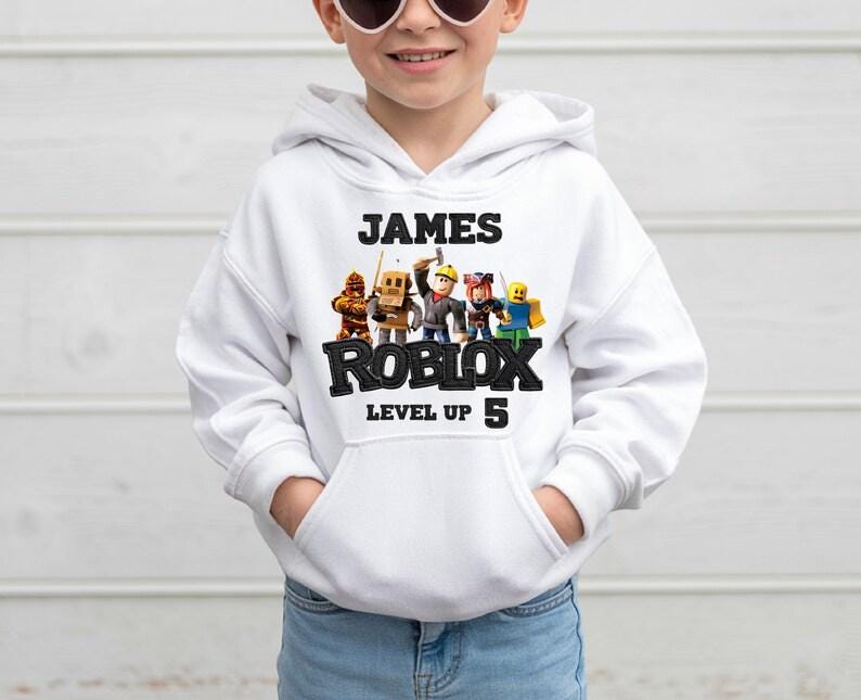 Puede incluir: Sudadera con capucha blanca con el nombre "JAMES" encima de "ROBLOX" en negro. Debajo de "ROBLOX" est&aacute; "LEVEL UP 5". La sudadera tiene un bolsillo delantero y un gr&aacute;fico de personajes de Roblox.