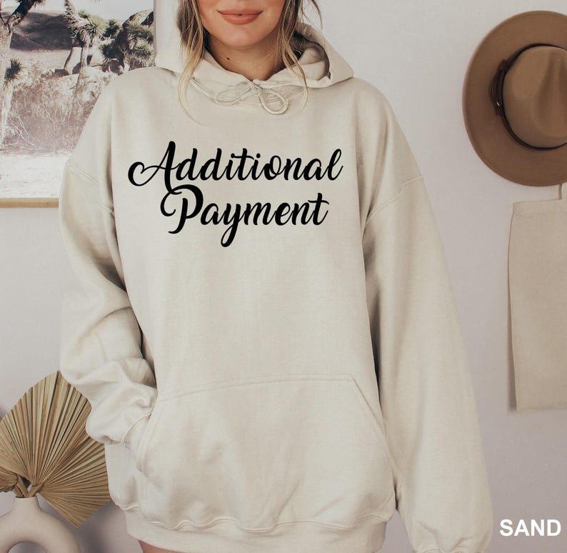 May include: A sand-coloured hoodie with the words "Additional Payment" in black script across the front. The hoodie has a drawstring hood and a front pocket. A hat hangs in the background.