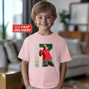 Puede incluir: Camiseta rosa claro con un gr&aacute;fico de un jugador de f&uacute;tbol con uniforme rojo. El n&uacute;mero 7 es visible. La palabra "RONALDO" est&aacute; impresa verticalmente en verde. Una pegatina de "FAST DELIVERY" es visible.