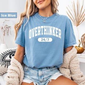 May include: Ice blue t-shirt with the word "OVERTHINKER" in large white block letters. Below the word is the text "24/7" in a white oval. The shirt is a short-sleeved crew neck.