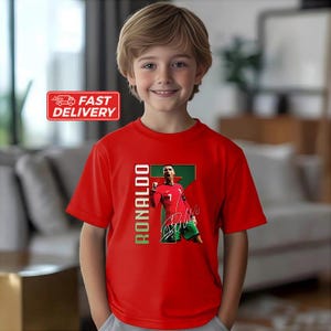 Puede incluir: Una camiseta roja con un retrato de Cristiano Ronaldo con uniforme de f&uacute;tbol. La camiseta tiene el nombre "RONALDO" en texto vertical verde y una firma. La camiseta la lleva un joven.
