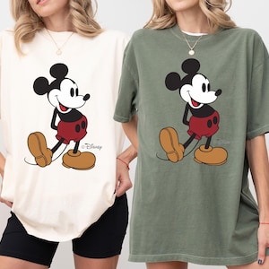 May include: Two oversized t-shirts, one ivory and one moss green, each featuring a large graphic of Mickey Mouse. The cartoon character is depicted in his classic outfit with red shorts, yellow shoes, and black gloves.