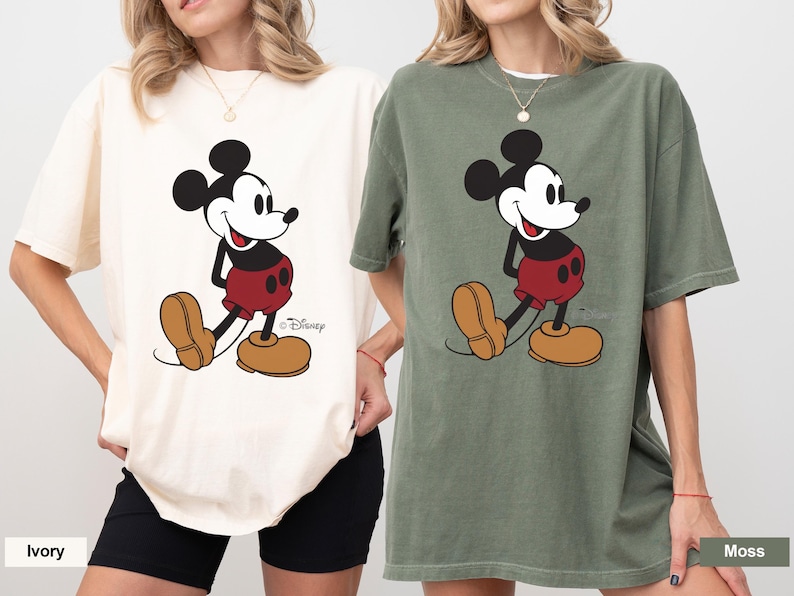 May include: Two oversized t-shirts, one ivory and one moss green, each featuring a large graphic of Mickey Mouse. The cartoon character is depicted in his classic outfit with red shorts, yellow shoes, and black gloves.