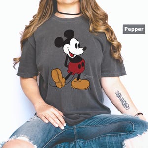 May include: A charcoal gray t-shirt featuring a classic Mickey Mouse graphic. Mickey is depicted in his iconic pose, wearing red shorts and yellow shoes. The Disney logo is visible. The shirt is labeled "Pepper".