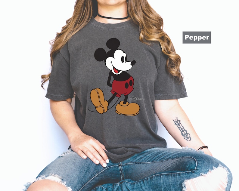 May include: A charcoal gray t-shirt featuring a classic Mickey Mouse graphic. Mickey is depicted in his iconic pose, wearing red shorts and yellow shoes. The Disney logo is visible. The shirt is labeled "Pepper".