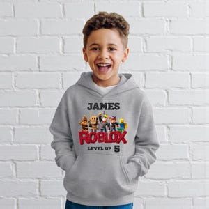 Puede incluir: Sudadera con capucha gris con el nombre "JAMES" sobre un gr&aacute;fico colorido de Roblox. El logotipo de Roblox es rojo, con "LEVEL UP 5" debajo. La sudadera la lleva un ni&ntilde;o sonriente.