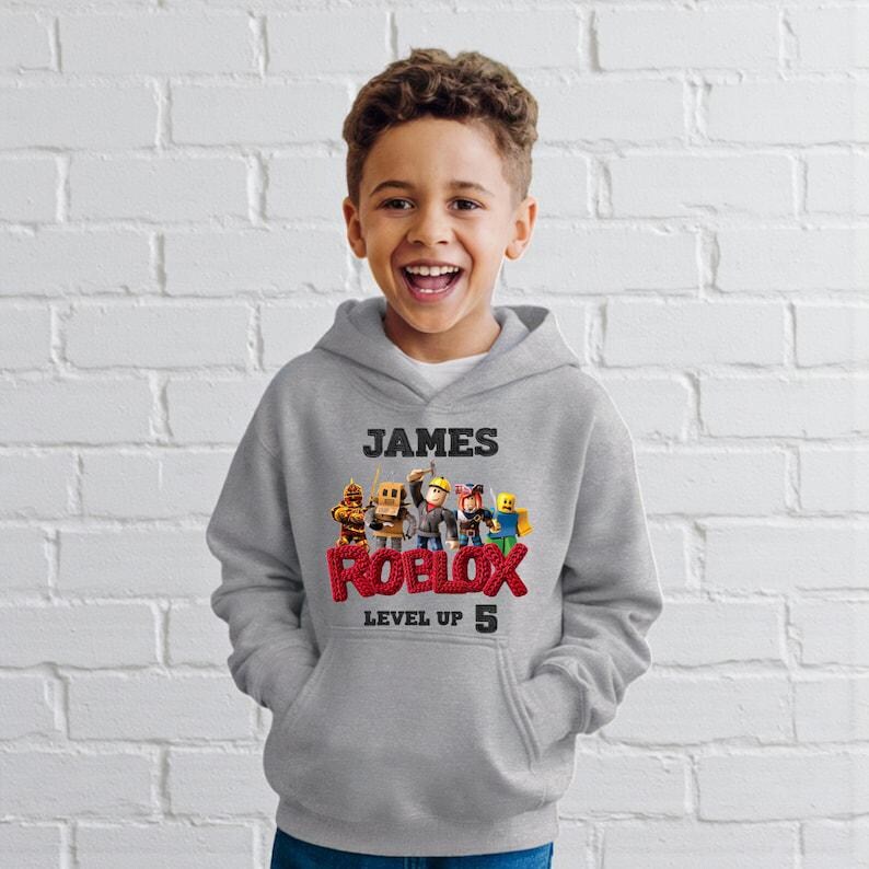 Puede incluir: Sudadera con capucha gris con el nombre "JAMES" sobre un gr&aacute;fico colorido de Roblox. El logotipo de Roblox es rojo, con "LEVEL UP 5" debajo. La sudadera la lleva un ni&ntilde;o sonriente.