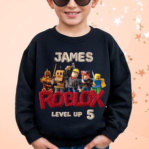 Puede incluir: Sudadera negra con el nombre "JAMES" sobre un logotipo rojo de "ROBLOX" y personajes de dibujos animados. Debajo del logotipo est&aacute;n las palabras "LEVEL UP" y el n&uacute;mero "5". Ropa para ni&ntilde;os.