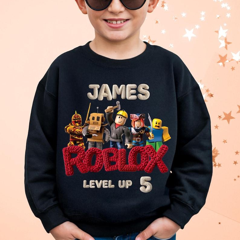 Puede incluir: Sudadera negra con el nombre "JAMES" sobre un logotipo rojo de "ROBLOX" y personajes de dibujos animados. Debajo del logotipo est&aacute;n las palabras "LEVEL UP" y el n&uacute;mero "5". Ropa para ni&ntilde;os.