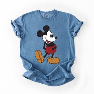May include: A blue t-shirt featuring a graphic of Mickey Mouse. Mickey is depicted in his classic attire: black ears, a white face, red shorts, and yellow shoes. The Disney logo is visible on the shirt.