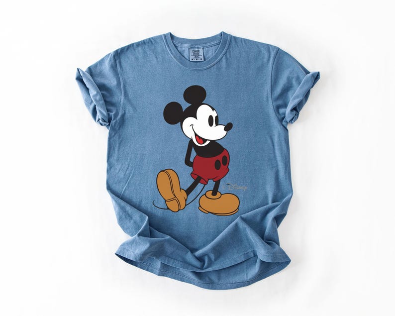 May include: A blue t-shirt featuring a graphic of Mickey Mouse. Mickey is depicted in his classic attire: black ears, a white face, red shorts, and yellow shoes. The Disney logo is visible on the shirt.